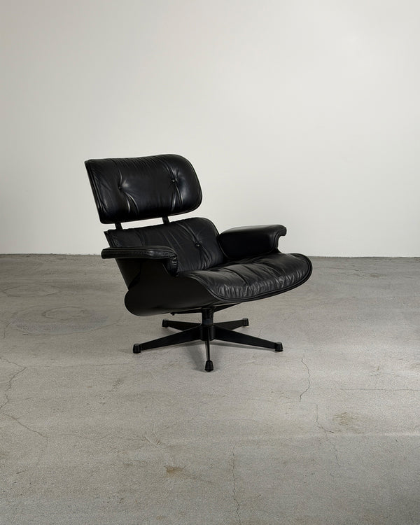 A classic black leather Eames-style lounge chair with a tufted headrest and five-star base, photographed in a minimalist studio setting with a concrete floor and plain wall.