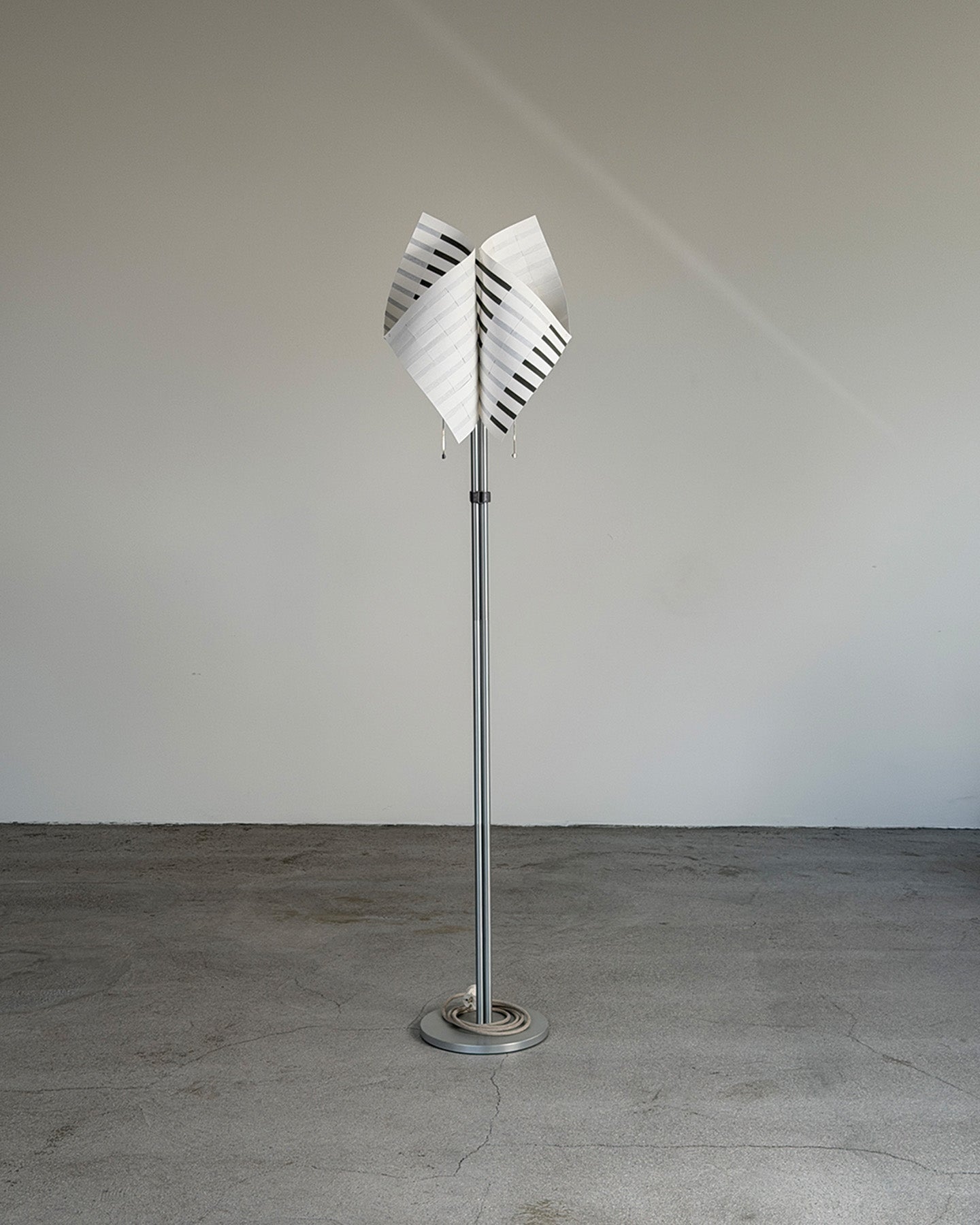 A modern TEX001 Studio D-ja floor lamp with a tall silver metal pole and a white shade featuring black diagonal stripes, standing on a circular base against a plain gray wall and concrete floor.
