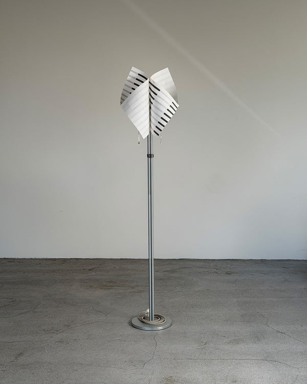 A modern TEX001 Studio D-ja floor lamp with a tall silver metal pole and a white shade featuring black diagonal stripes, standing on a circular base against a plain gray wall and concrete floor.