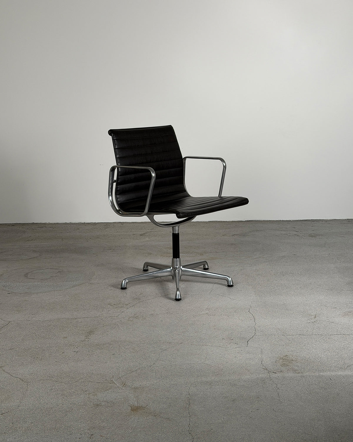 A modern black leather swivel office chair with tubular armrests and a chrome five-star base, set against a minimalist gray concrete floor and wall.