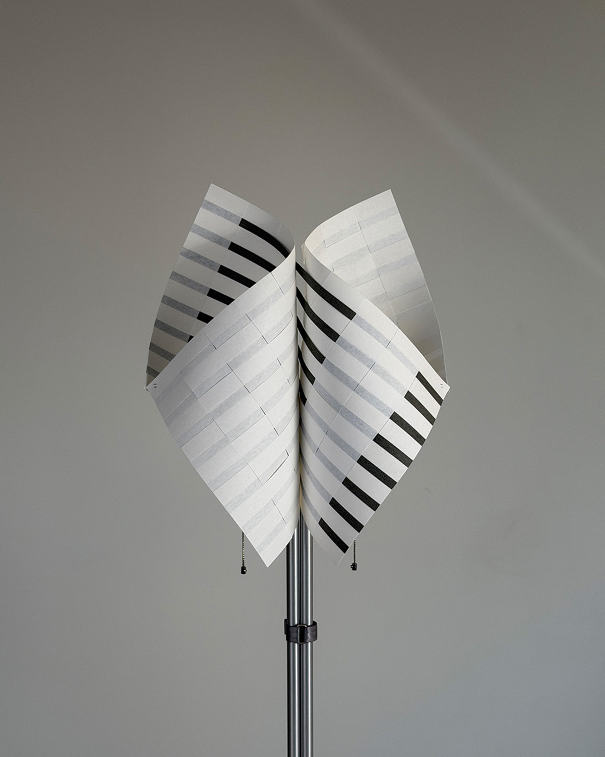 A modern sculptural floor lamp featuring two large, curved white panels with black horizontal stripes, mounted on a slim metallic pole against a neutral gray background.