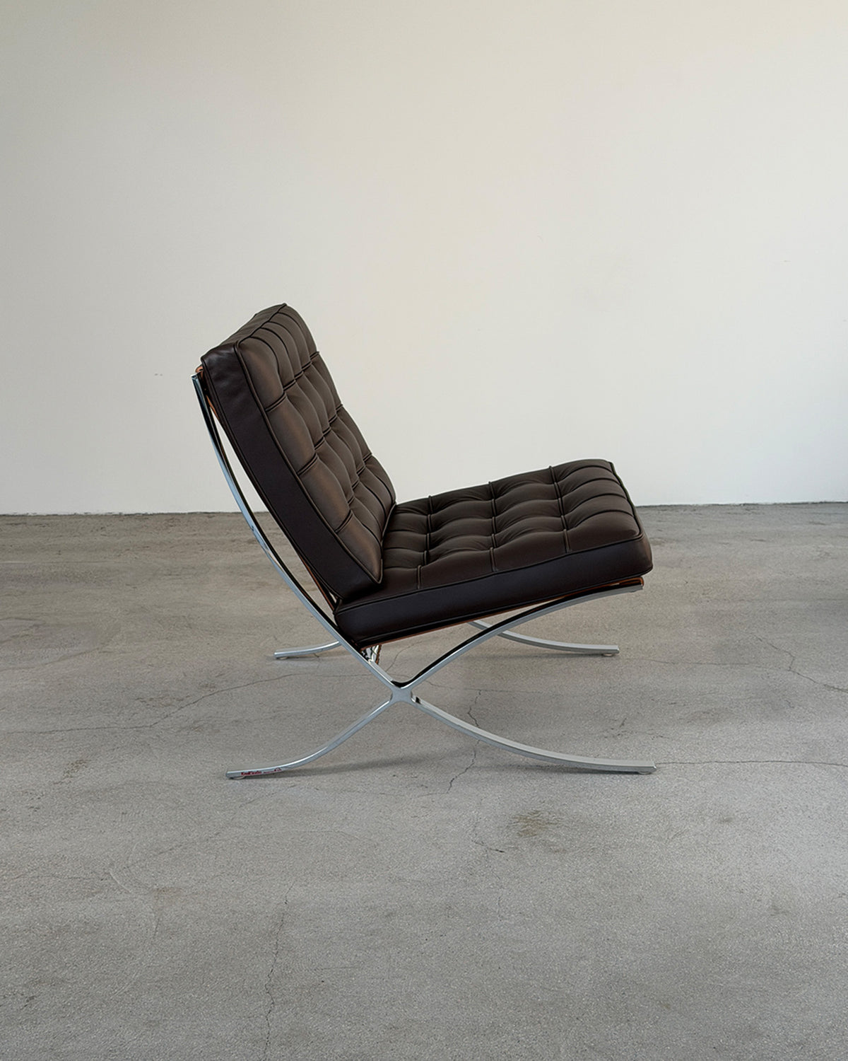 A modern Barcelona lounge chair with dark brown tufted leather upholstery and a chrome steel X-frame base, photographed in a minimalist studio setting with a concrete floor and plain off-white wall.