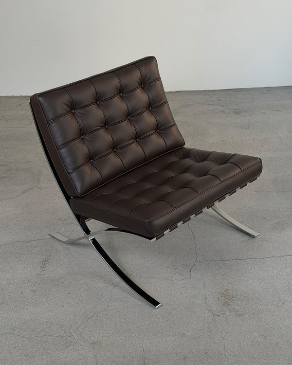 A modern brown leather armchair with a tufted backrest and seat, supported by a chrome S-shaped base, photographed in a minimalist studio setting with a concrete floor and plain wall.
