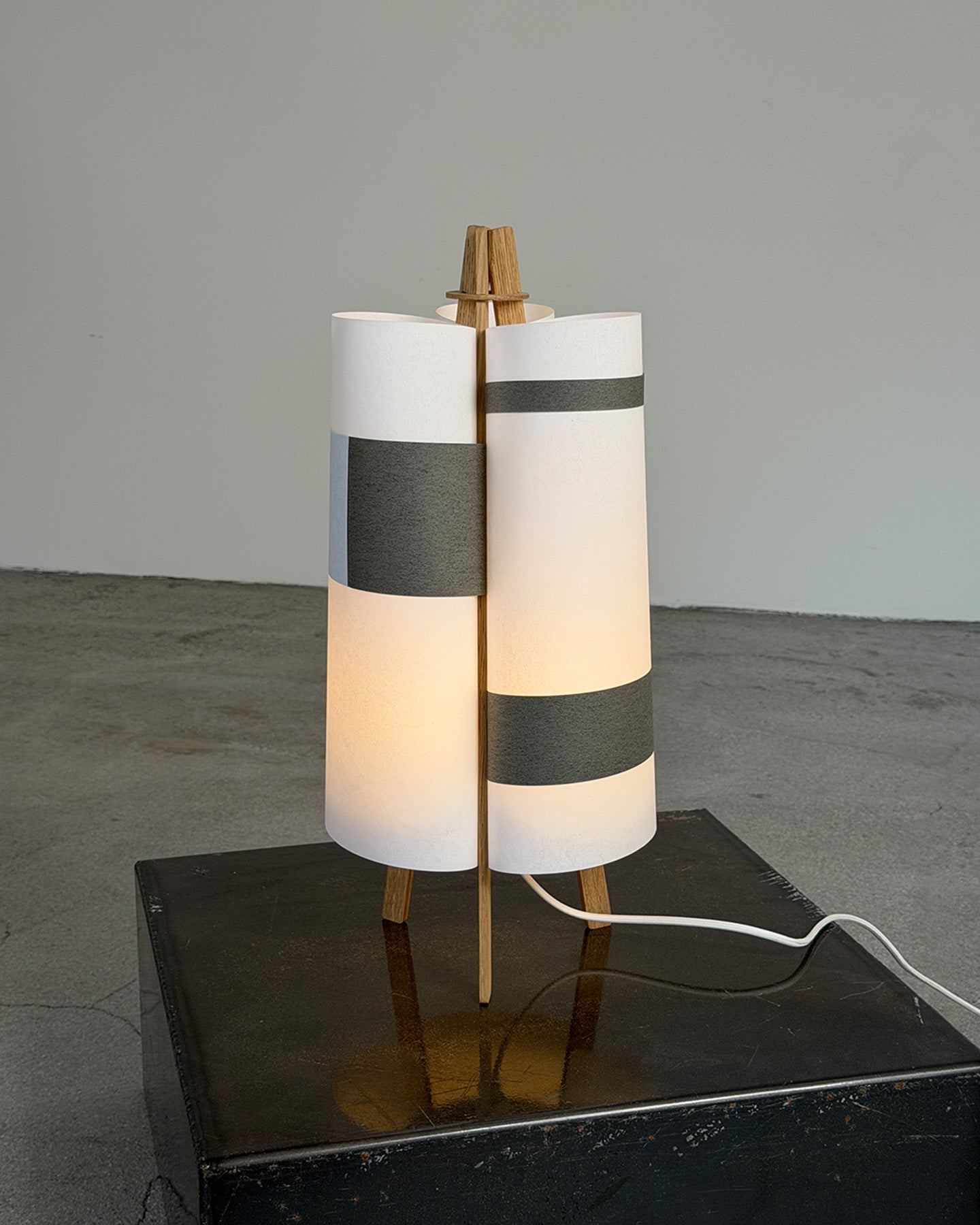 A modern table lamp with a cylindrical white shade featuring horizontal gray bands, illuminated with a warm glow, supported by a light wood frame and standing on a black base.