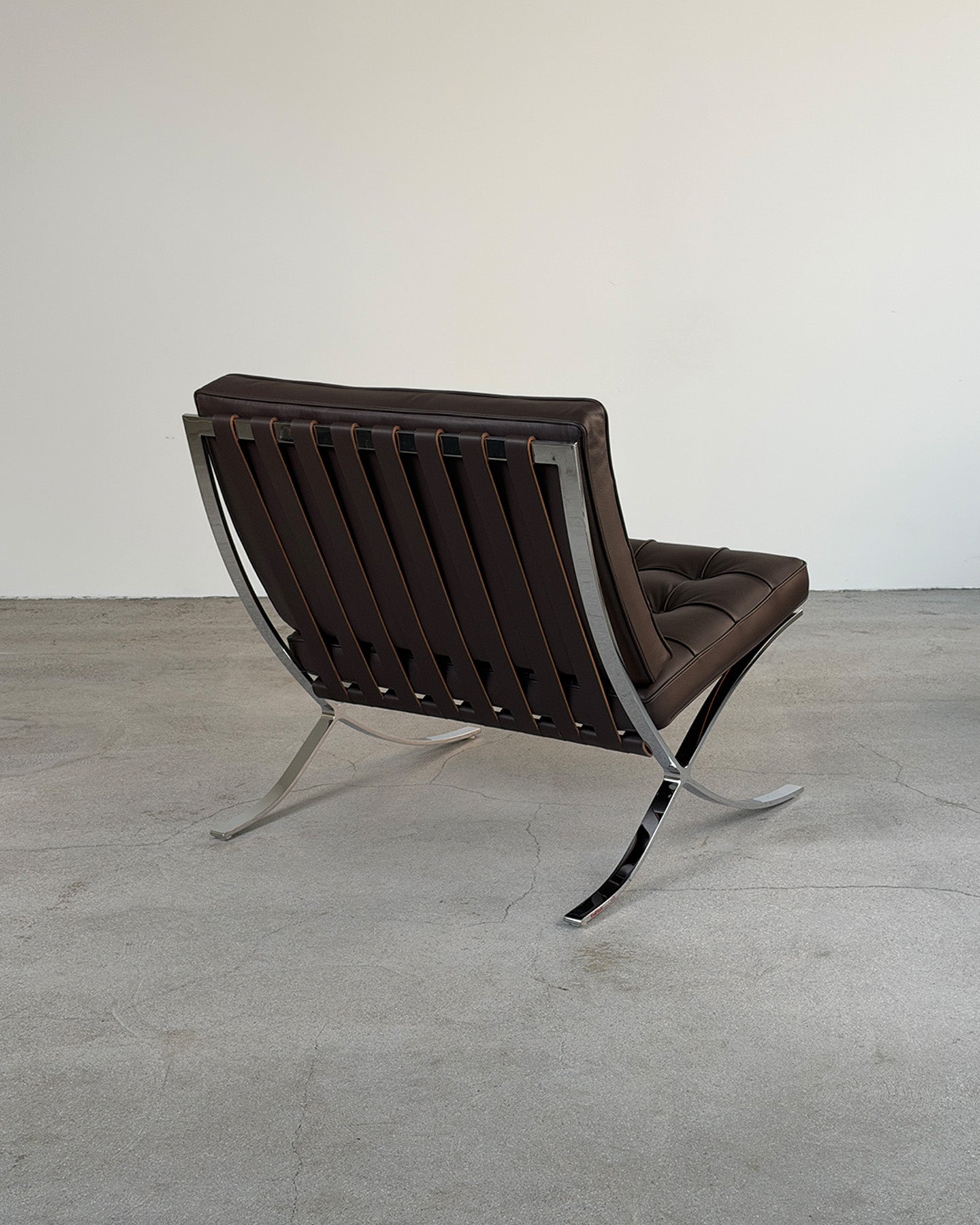 A mid-century modern Barcelona-style lounge chair with dark brown leather slatted back and seat, supported by a polished chrome metal frame, photographed in a minimalist studio setting with a concrete floor and plain off-white wall.