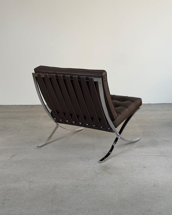 A mid-century modern Barcelona-style lounge chair with dark brown leather slatted back and seat, supported by a polished chrome metal frame, photographed in a minimalist studio setting with a concrete floor and plain off-white wall.