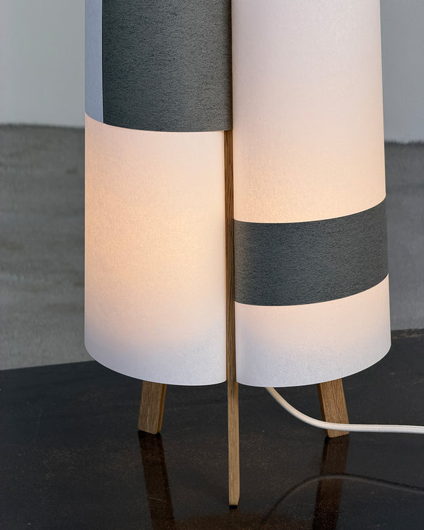 A modern table lamp with two illuminated white cylindrical shades, dark gray textured horizontal bands, and light wood legs on a dark reflective surface.