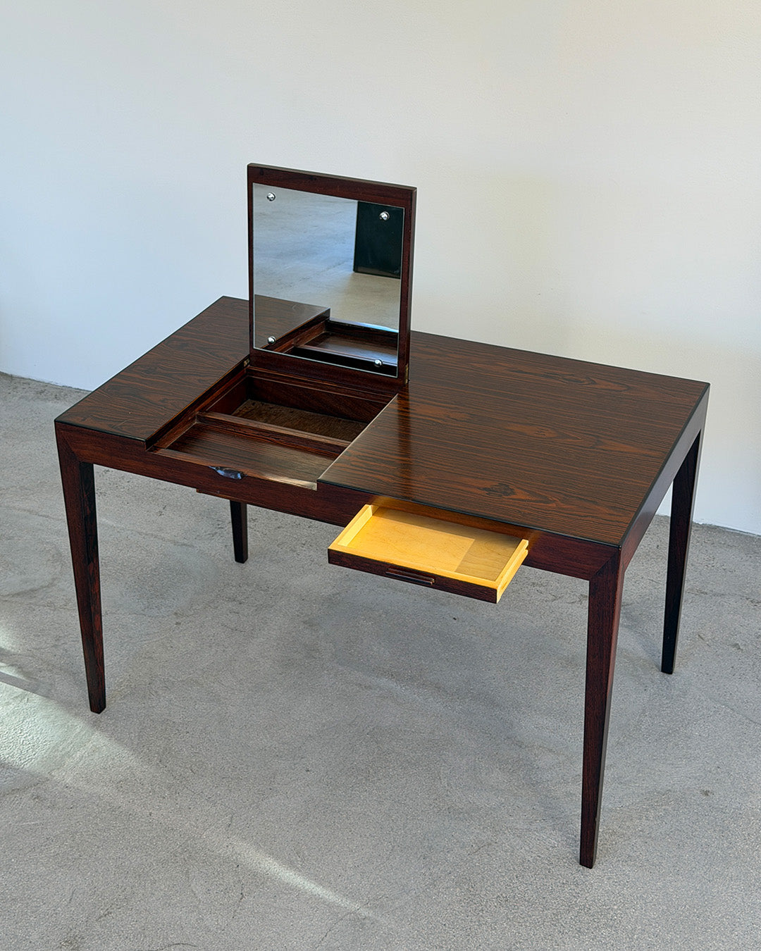 Wooden vanity table with mirror and drawer on a plain floor.