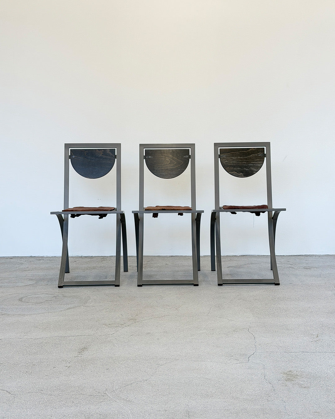 Three metal chairs with wooden seats on a light wooden floor against a white wall.