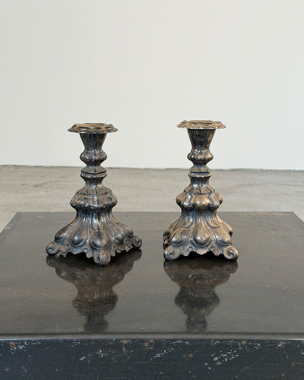 A pair of ornate antique-style metal candlesticks with sculpted floral bases, displayed side by side on a dark reflective surface against a plain light gray wall.