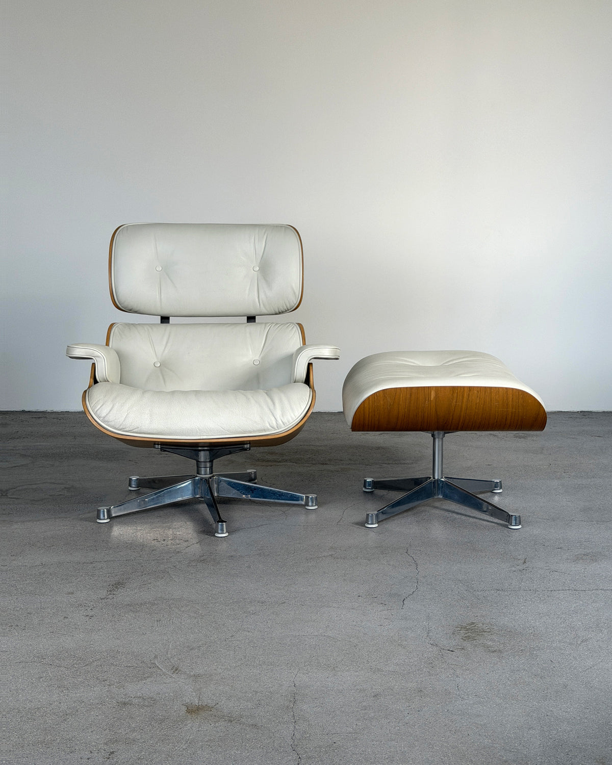 A mid-century modern lounge chair with a matching ottoman, featuring off-white leather upholstery, a warm wood frame, and chrome star bases, set against a plain light gray wall and concrete floor.